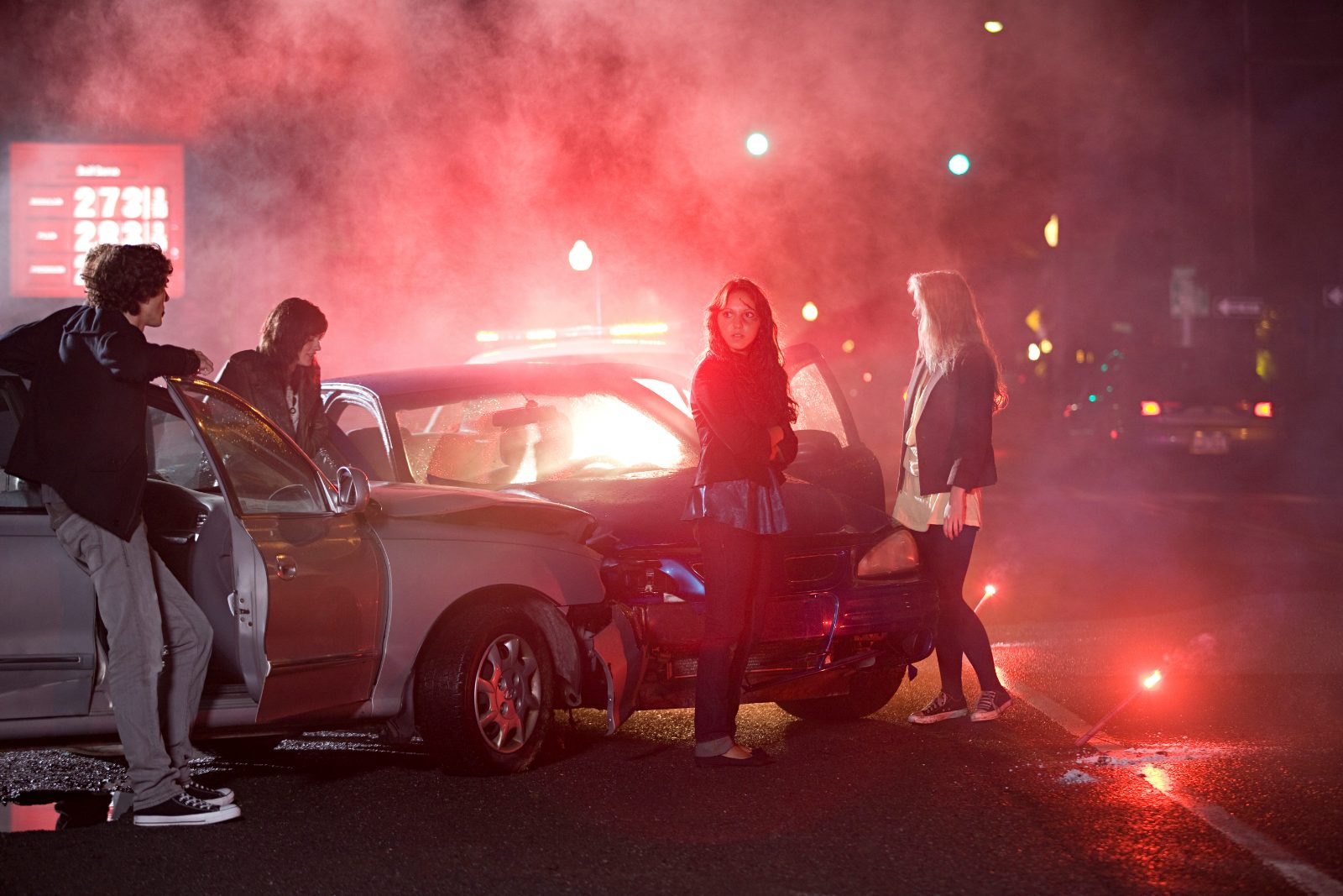 Teenagers gathered at night around damaged cars and red smoke